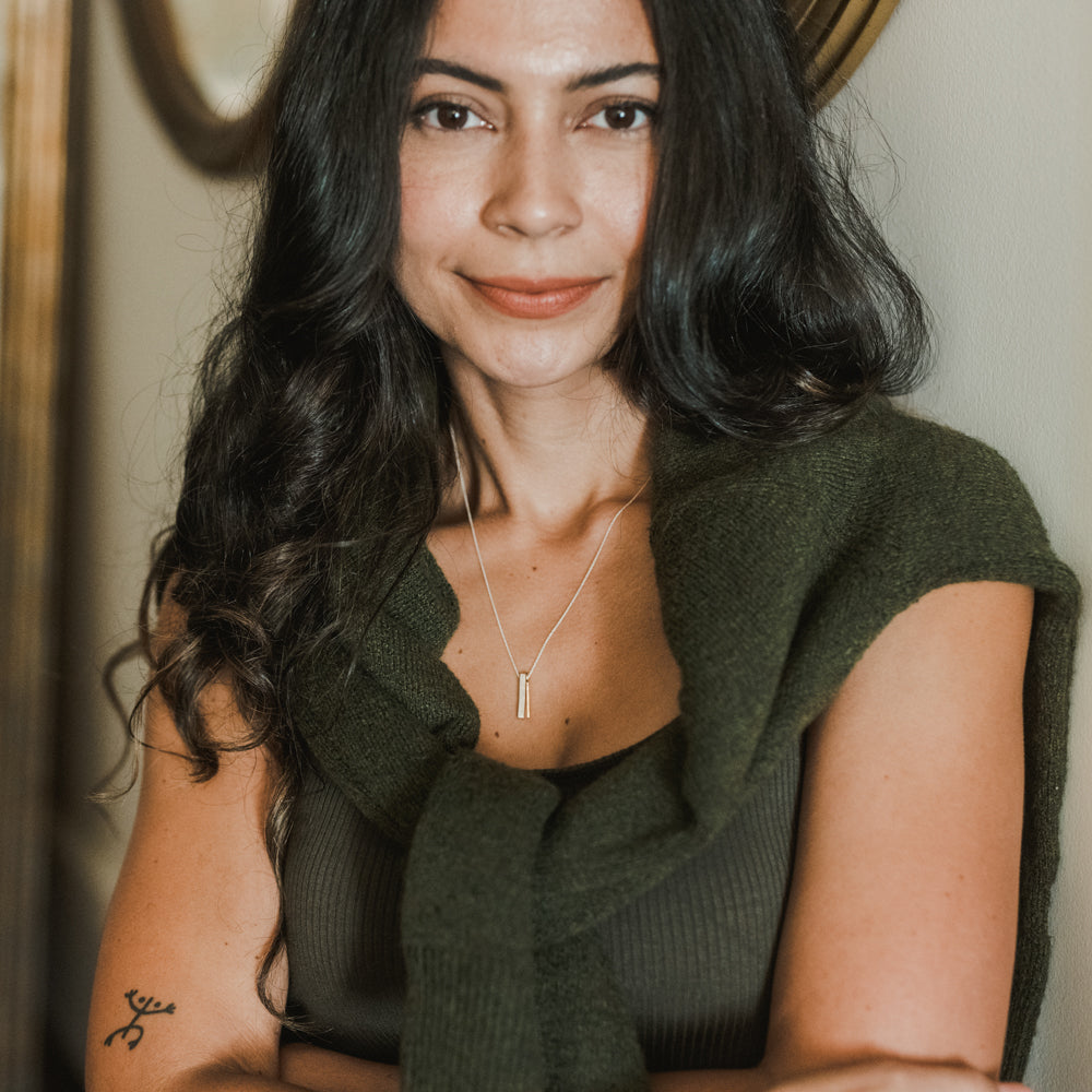 A woman with long dark hair and a green sweater draped over her shoulders stands with her arms crossed. She has a tattoo on her left arm and wears the best-selling "Through Thick & Thin Necklace" by Becoming Jewelry, making it the perfect gift to symbolize enduring bonds.