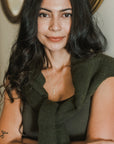 A woman with long dark hair and a green sweater draped over her shoulders stands with her arms crossed. She has a tattoo on her left arm and wears the best-selling "Through Thick & Thin Necklace" by Becoming Jewelry, making it the perfect gift to symbolize enduring bonds.