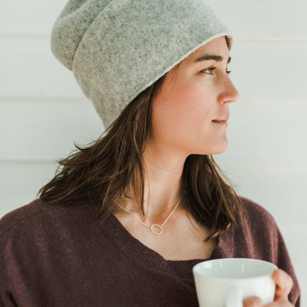 Woman in beanie and a Becoming Jewelry Karma Necklace holding a mug and looking to the side against a white background.