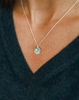 Close-up of a person wearing the Blossom Necklace by Becoming Jewelry, featuring a round pendant, against the backdrop of a dark V-neck shirt, symbolizing the growth and blossoming flowers that come with deep friendship.