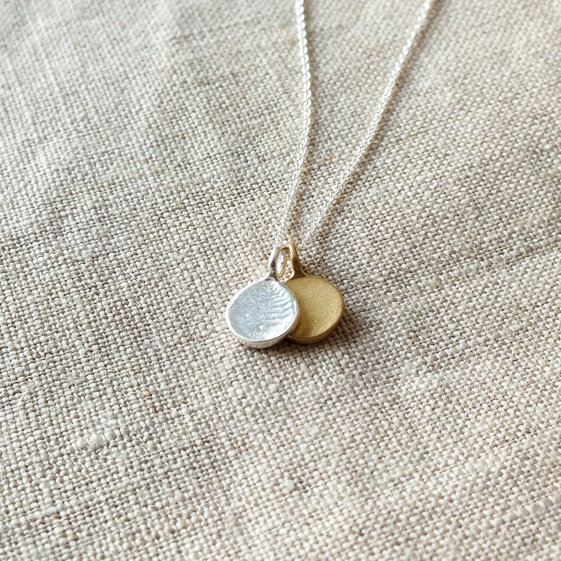 Close-up of the Count My Blessings Necklace by Becoming Jewelry, featuring two charming drop discs pendants—one in silver and one in gold—on a textured beige fabric background.