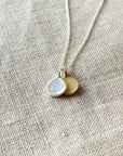 Close-up of the Count My Blessings Necklace by Becoming Jewelry, featuring two charming drop discs pendants—one in silver and one in gold—on a textured beige fabric background.
