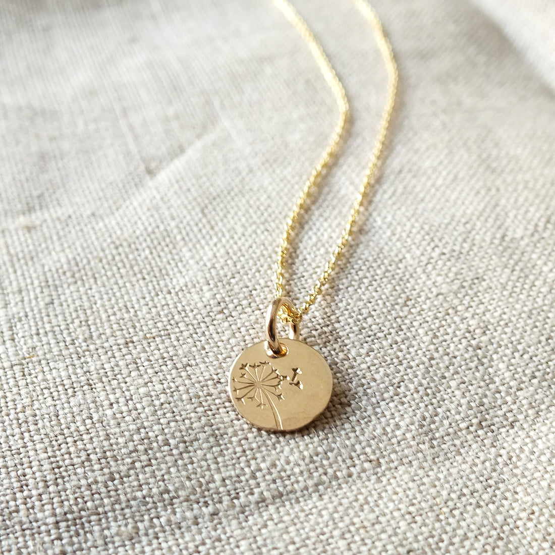 The Dandelion Wishes Necklace from Becoming Jewelry, featuring a gold pendant with a round charm engraved with a dandelion design and inspirational words, is placed on a textured fabric surface.