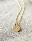The Dandelion Wishes Necklace from Becoming Jewelry, featuring a gold pendant with a round charm engraved with a dandelion design and inspirational words, is placed on a textured fabric surface.