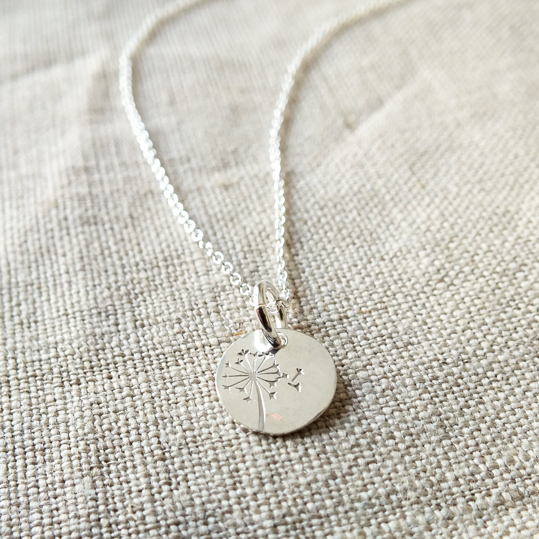 The Dandelion Wishes Necklace by Becoming Jewelry showcases a round pendant engraved with a dandelion charm, elegantly displayed on a textured beige fabric background. This sophisticated piece is presented on a beautifully designed necklace card featuring inspirational words.