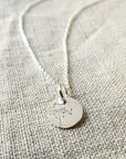 The Dandelion Wishes Necklace by Becoming Jewelry showcases a round pendant engraved with a dandelion charm, elegantly displayed on a textured beige fabric background. This sophisticated piece is presented on a beautifully designed necklace card featuring inspirational words.