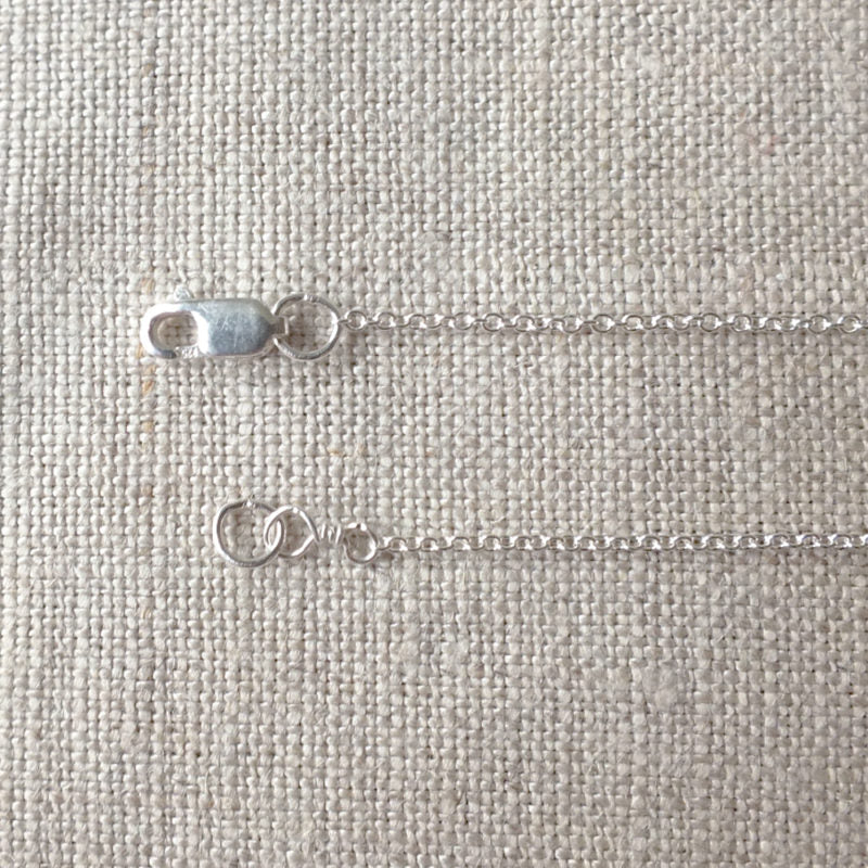 A close-up of the Replacement Chain, Silver by Becoming Jewelry featuring a lobster clasp on a textured beige surface.