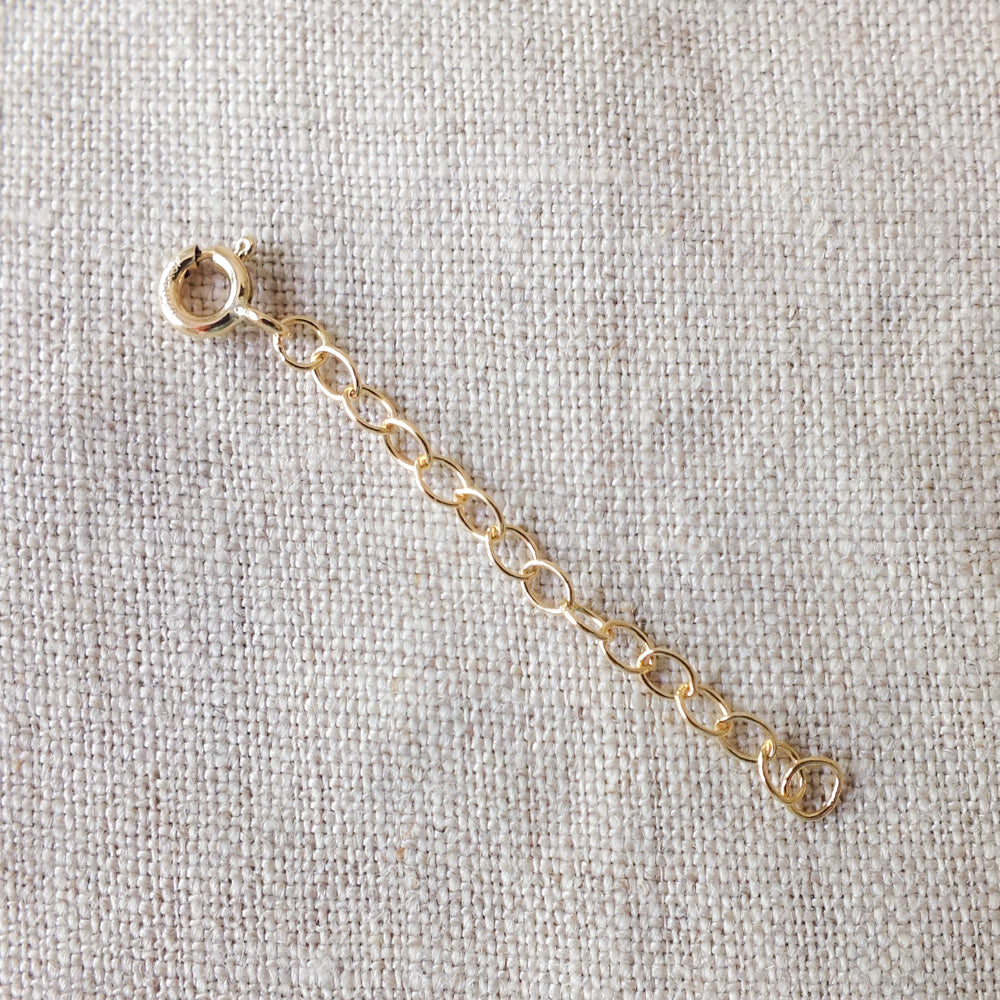 A short gold Chain Extender by Becoming Jewelry, featuring a round clasp and a subtle chain extender, resting on a textured beige fabric surface.