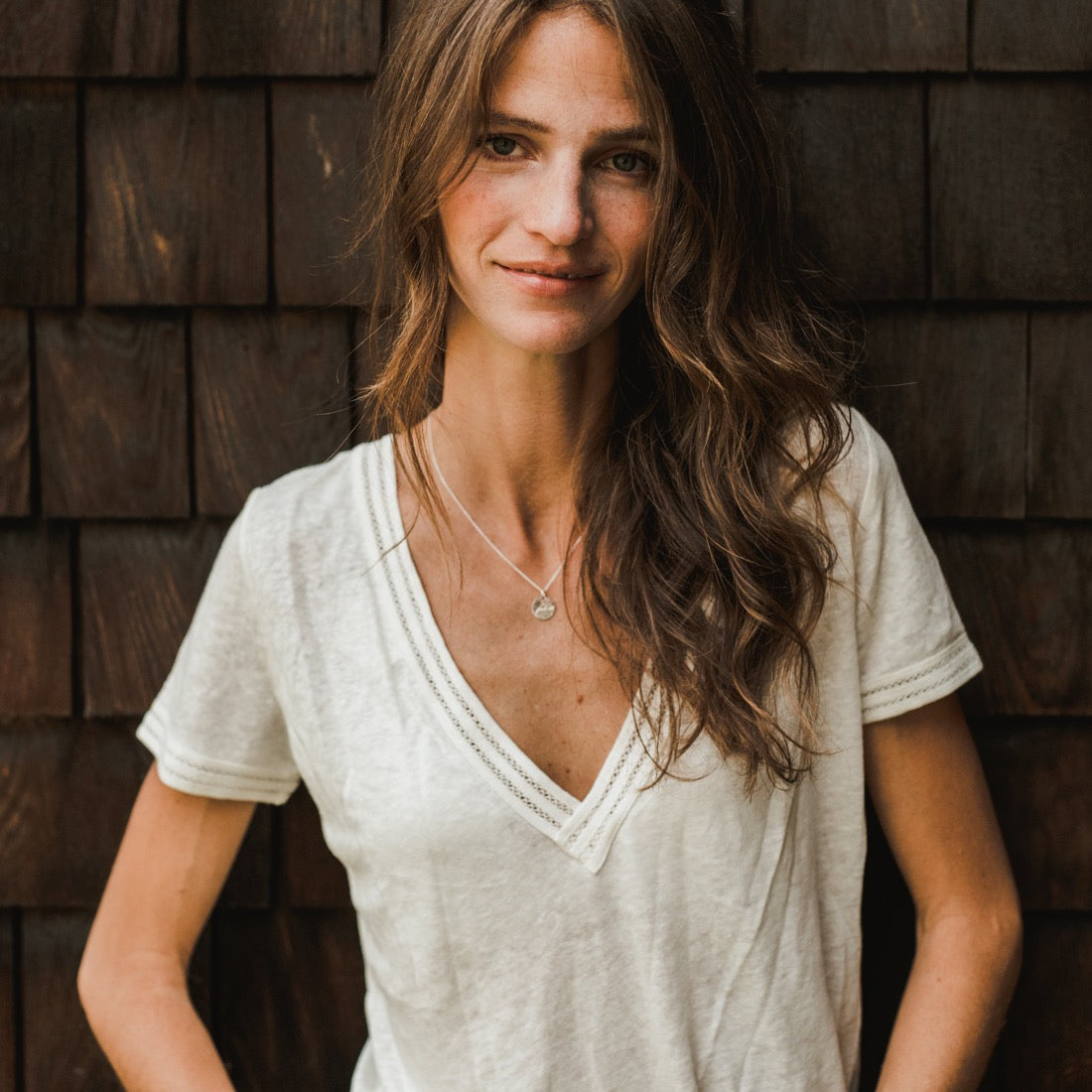 A woman with long wavy hair is wearing a white V-neck T-shirt and the "Fuck It Necklace" from Becoming Jewelry, standing in front of a wooden shingle wall.