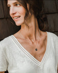 A person with long hair wearing a white V-neck shirt and the Fuck It Necklace by Becoming Jewelry, which features a small round charm, stands against a wooden background, looking to the side and smiling.
