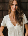 A woman with long wavy hair is wearing a white V-neck T-shirt and the "Fuck It Necklace" from Becoming Jewelry, standing in front of a wooden shingle wall.