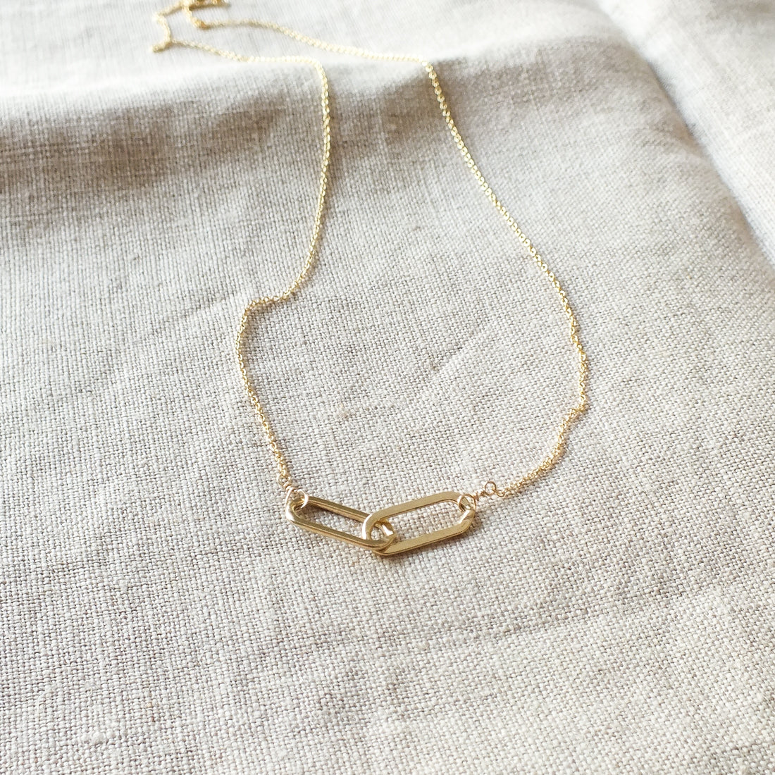A delicate gold chain necklace with a centerpiece featuring two interlocking links, displayed on a light fabric background. Perfect as a gift, this Linked Together Necklace from Becoming Jewelry symbolizes an unbreakable connection.