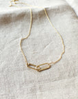 A delicate gold chain necklace with a centerpiece featuring two interlocking links, displayed on a light fabric background. Perfect as a gift, this Linked Together Necklace from Becoming Jewelry symbolizes an unbreakable connection.