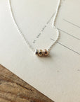 The Three Things Necklace by Becoming Jewelry, featuring a delicate chain adorned with three metallic beads in silver, gold, and copper, rests gracefully on a piece of text-covered paper. The paper is placed on a wooden surface, adding an aura of love to the scene.