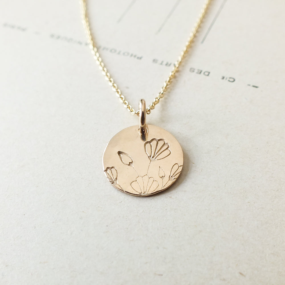 The Blossom Necklace by Becoming Jewelry features a gold disc pendant engraved with a floral design of blossoming flowers, elegantly hanging from a delicate gold chain and beautifully showcased on a light-colored background.