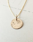 The Blossom Necklace by Becoming Jewelry features a gold disc pendant engraved with a floral design of blossoming flowers, elegantly hanging from a delicate gold chain and beautifully showcased on a light-colored background.