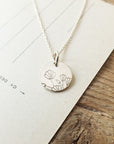 The Blossom Necklace by Becoming Jewelry, a silver necklace with a round pendant featuring an engraved blossom design, rests on a piece of paper on a wooden surface, symbolizing growth and friendship.