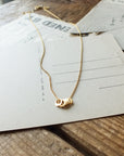 A Blessings Necklace by Becoming Jewelry with a pendant resting on a postcard on a wooden surface.