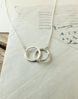 A silver Joined for Life Necklace from Becoming Jewelry, featuring two interlocking rings, rests on an old letter with handwritten text, symbolizing an unbreakable bond.