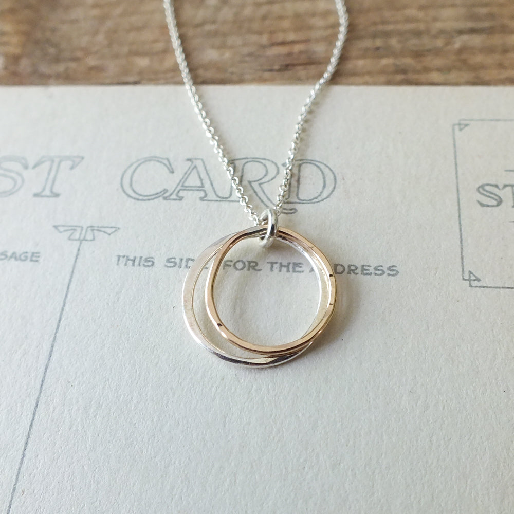 The Friendship Circles Necklace by Becoming Jewelry, featuring interlocking mixed metal pendants, is beautifully displayed on a postcard background.