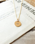 The Heart Necklace from Becoming Jewelry, featuring a round pendant with a small heart engraving, is elegantly placed on a textured surface next to a piece of paper, making it a meaningful gift.