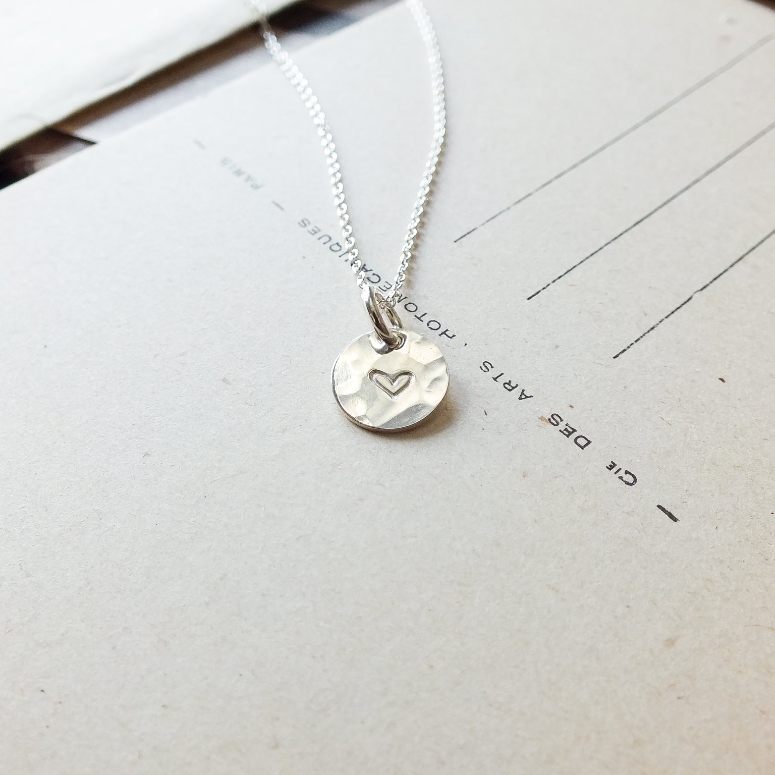 A Becoming Jewelry sterling silver Heart Necklace on a chain, resting on a piece of paper with printed text.