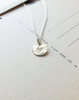 A silver Heart Necklace from Becoming Jewelry, featuring a circular pendant with a small heart design, rests on a beige postcard, making for a meaningful gift.