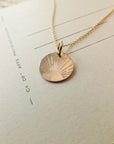 A Becoming Jewelry Irish Blessing Necklace with a round gold pendant featuring a sunburst design lies on a piece of paper with printed text, making it perfect for gifting.