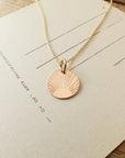 A gold *Becoming Jewelry* Irish Blessing Necklace with a round pendant featuring a sunburst design, placed on a piece of paper with faint text.