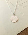 The Irish Blessing Necklace by Becoming Jewelry, a silver necklace featuring a round pendant with an engraved sunburst design, ideal for gifting, is elegantly placed on beige paper with partially visible text.