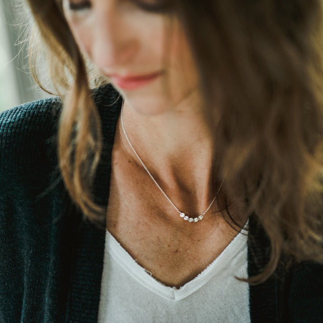 Close-up of a person wearing a black cardigan, white shirt, and a silver Mothers & Daughters Necklace from Becoming Jewelry with multiple small beads, looking down slightly.