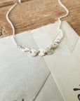 The Mothers & Daughters Necklace by Becoming Jewelry, a silver chain with delicate circular links symbolizing connection, rests elegantly on top of an envelope on a wooden surface.