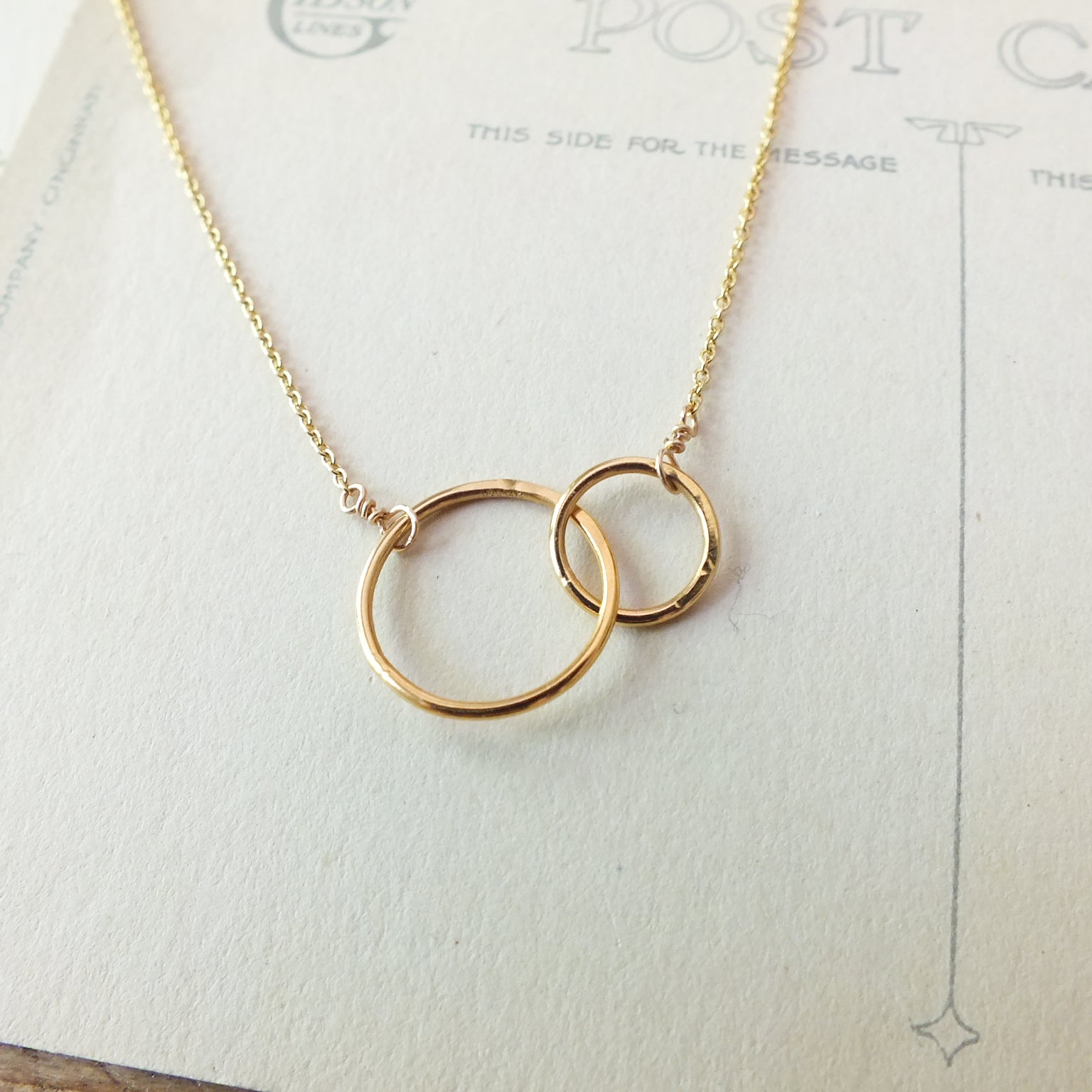 Mother Necklace by Becoming Jewelry, featuring two interlocking rings that symbolize a mother's love, elegantly displayed on a vintage postcard background.