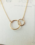 Mother Necklace by Becoming Jewelry, featuring two interlocking rings that symbolize a mother's love, elegantly displayed on a vintage postcard background.