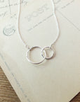 The Mother Necklace by Becoming Jewelry features interlocking silver circles on a minimalist chain, set against a vintage postcard background to symbolize mother's love and the special relationship.