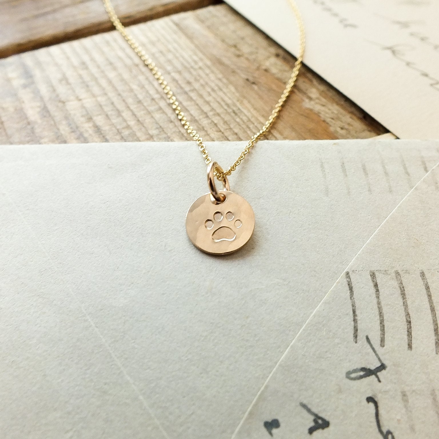 A Paw Print Necklace by Becoming Jewelry, featuring a gold chain with a circular pendant and stamped paw print charm, rests on top of a white envelope and papers.