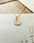 A Paw Print Necklace by Becoming Jewelry, featuring a gold chain with a circular pendant and stamped paw print charm, rests on top of a white envelope and papers.
