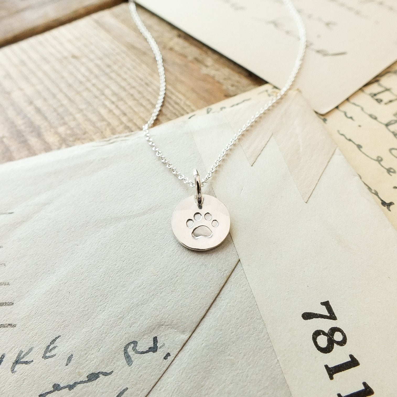 A silver Paw Print Necklace from Becoming Jewelry, featuring a circular pendant with a paw print charm, rests on top of three envelopes and letters, making it the perfect gift for animal lovers.