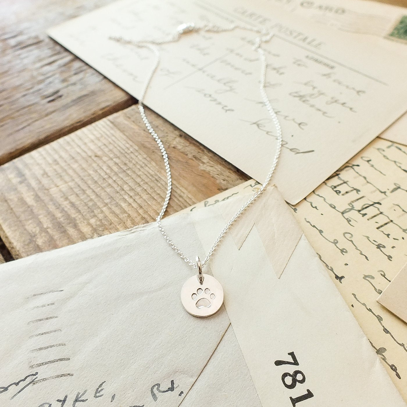 The "Paw Print Necklace" by Becoming Jewelry, featuring a round paw print charm in silver, rests on a pile of vintage letters and postcards on a wooden surface, making it the perfect gift for animal lovers.
