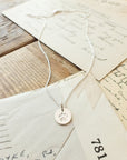 The "Paw Print Necklace" by Becoming Jewelry, featuring a round paw print charm in silver, rests on a pile of vintage letters and postcards on a wooden surface, making it the perfect gift for animal lovers.