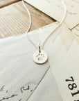 A silver Paw Print Necklace from Becoming Jewelry, featuring a circular pendant with a paw print charm, rests on top of three envelopes and letters, making it the perfect gift for animal lovers.