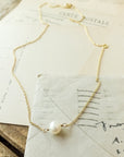 A delicate gold chain with a single pearl pendant, known as the Simple Pleasures Pearl Necklace by Becoming Jewelry, is laid on top of vintage postcards on a wooden surface.