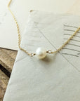 The Simple Pleasures Pearl Necklace by Becoming Jewelry, featuring a gold chain and a single pearl pendant, is displayed on top of a rustic wooden surface and envelopes, showcasing its timeless beauty.