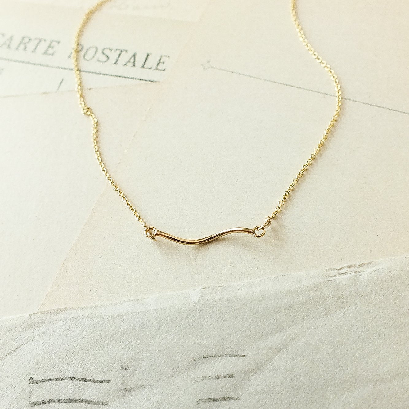The Path Necklace by Becoming Jewelry features a minimalist wave-shaped pendant in delicate gold, presented on a vintage-style paper background—a gentle reminder to always follow your heart.