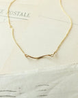 The Path Necklace by Becoming Jewelry features a minimalist wave-shaped pendant in delicate gold, presented on a vintage-style paper background—a gentle reminder to always follow your heart.
