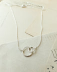 Sisters Necklace by Becoming Jewelry, featuring two intertwined circles, elegantly displayed on a stack of vintage postcards with handwritten text—an ideal gift for a beloved sister.