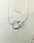 The Sisters Necklace by Becoming Jewelry, featuring two intertwined circular pendants, sits on a light beige background adorned with pieces of vintage paper, making it a perfect gift for a beloved sister.