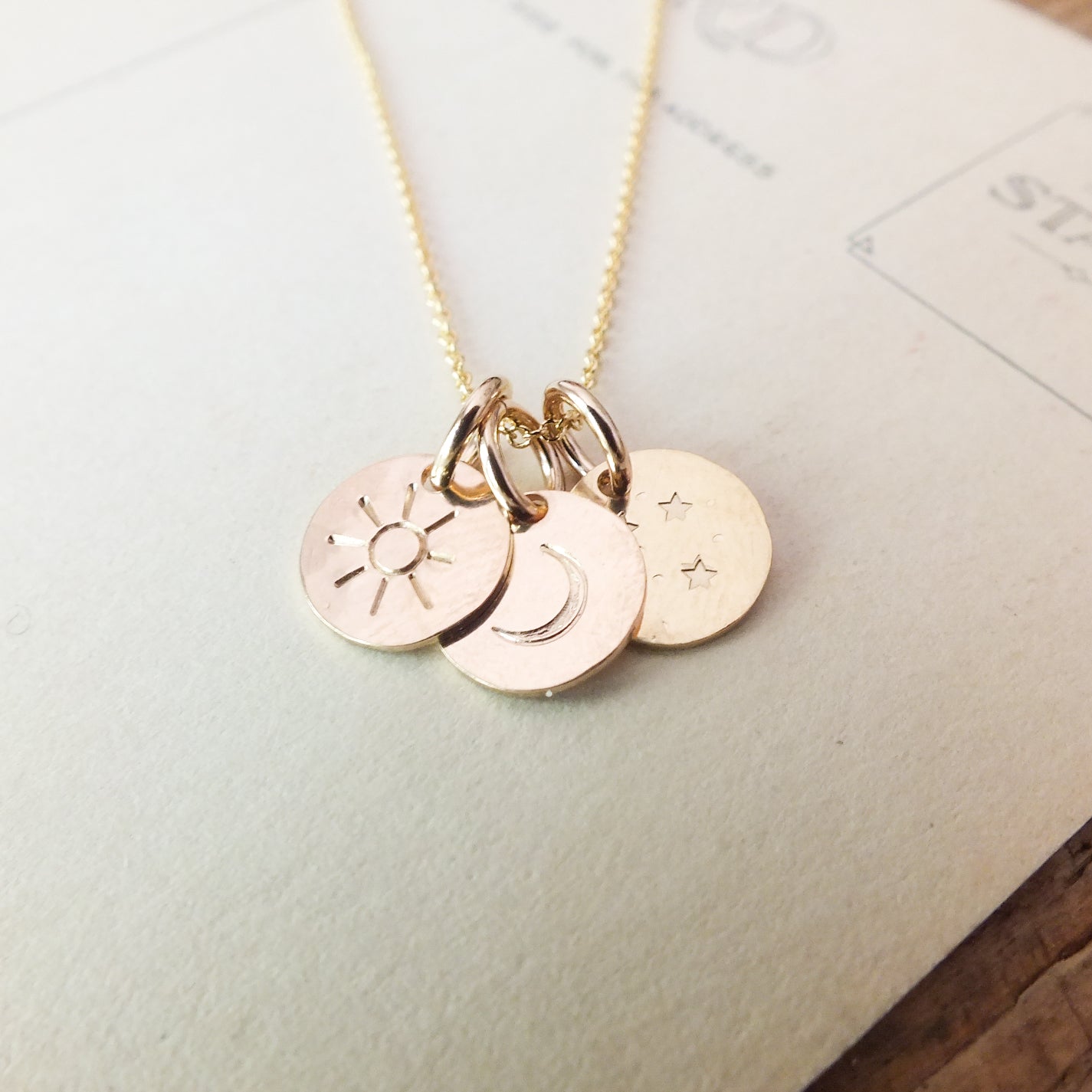 The Sun Moon and Stars Necklace by Becoming Jewelry features four small, round pendants engraved with a sun charm, a crescent moon, two stars, and another crescent moon. The necklace is beautifully displayed on a light-colored surface.
