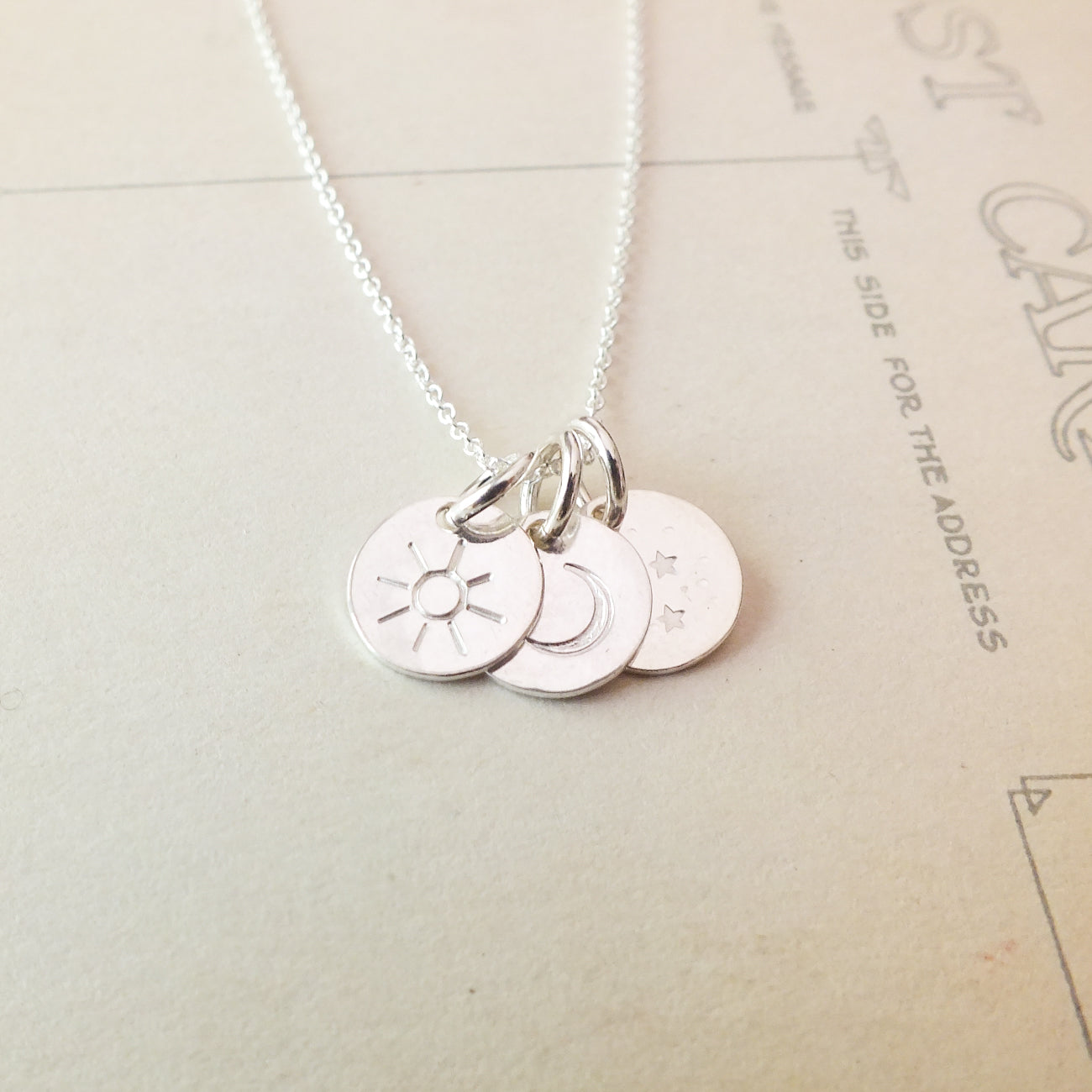 The Sun Moon and Stars Necklace by Becoming Jewelry is a stunning silver piece featuring three round pendants, each adorned with a unique symbol: a sun charm, a moon charm, and stars. This beautiful star gift is displayed on a light-colored surface with printed text in the background.
