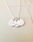 The Sun Moon and Stars Necklace by Becoming Jewelry is a stunning silver piece featuring three round pendants, each adorned with a unique symbol: a sun charm, a moon charm, and stars. This beautiful star gift is displayed on a light-colored surface with printed text in the background.
